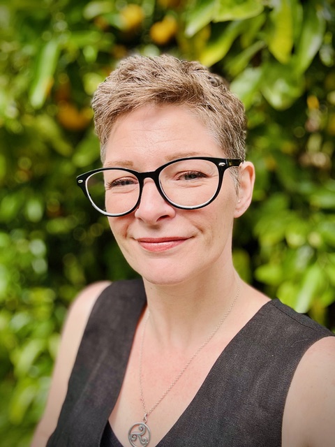Head and shoulders of a Caucasian person standing in front of a leafy background. They have very short hair and wear black-framed spectacles and a black vest. They are looking at the camera smiling slightly with their mouth closed. 